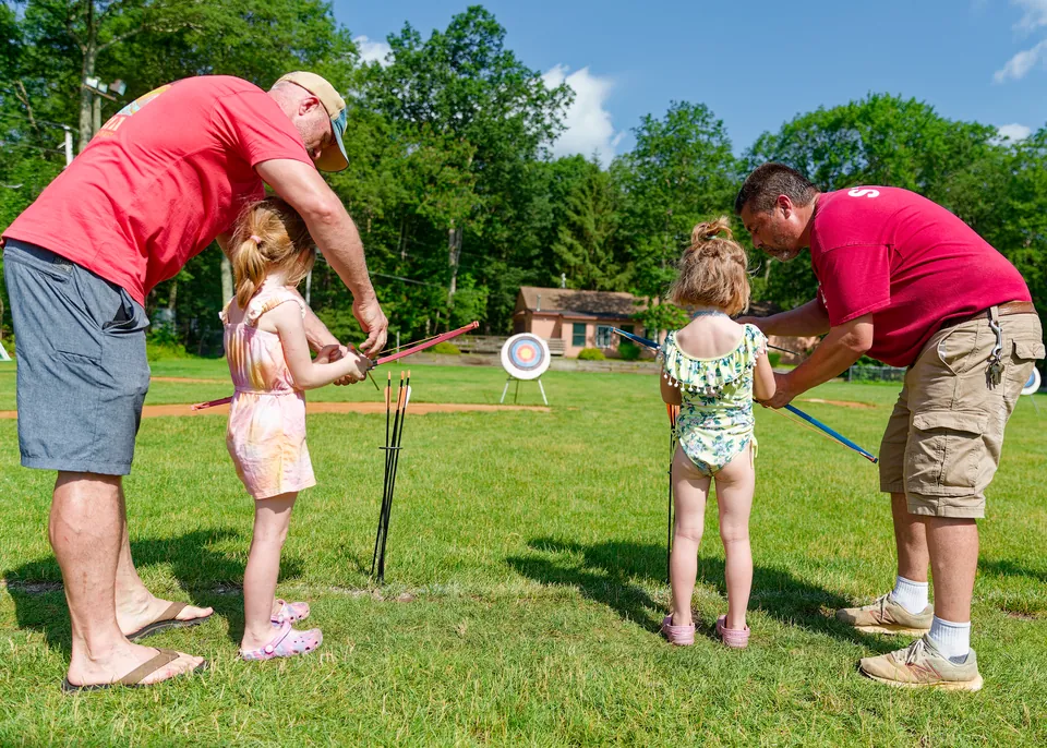 Two dads helping their daughters with archery on the lawn