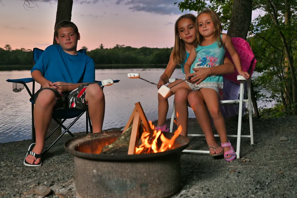 Three kids toasting marshmallows at a campfire by the lake at dusk